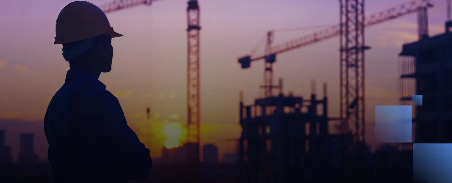 Silhouette of a construction worker wearing a helmet with cranes and buildings at sunset.