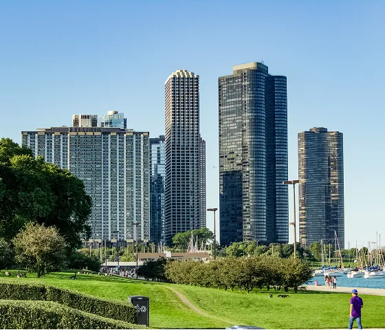 Green park area with trees and grass in front of tall modern skyscrapers near water with boats.