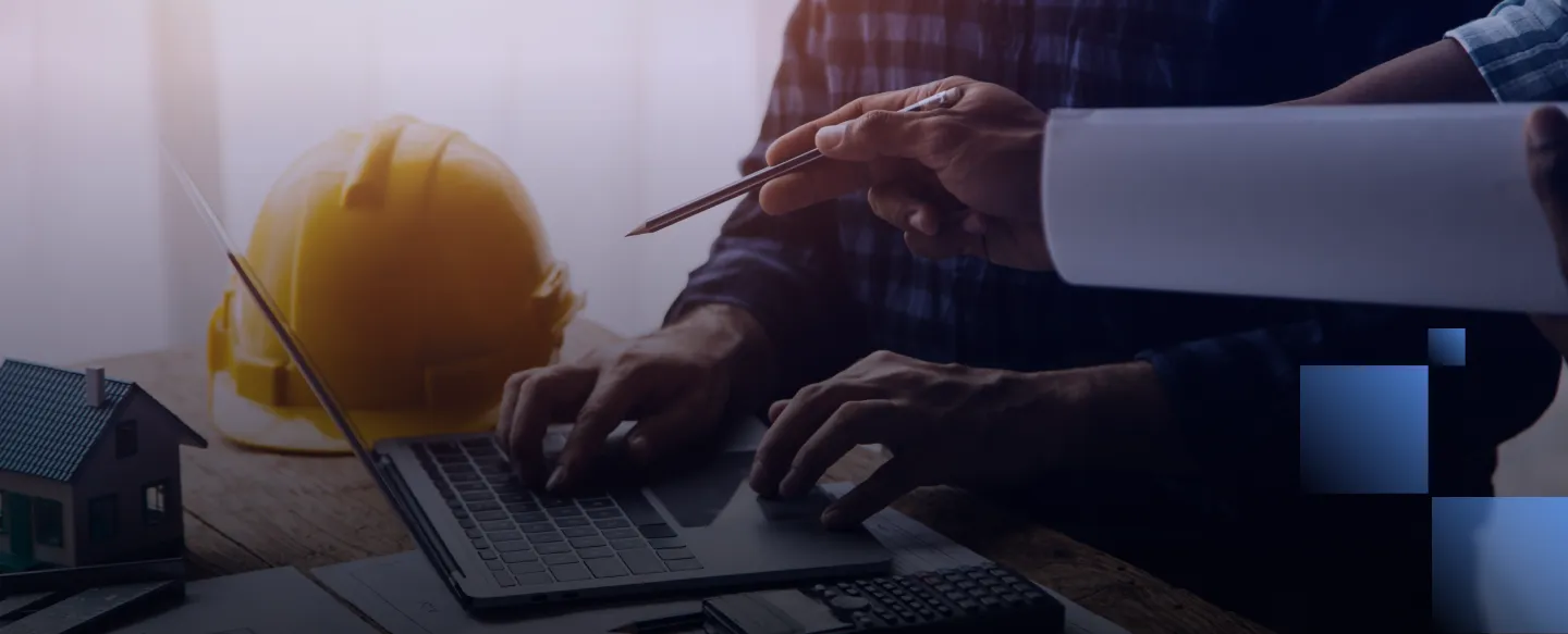 Two people working at a laptop with a yellow safety helmet and house model on the table.