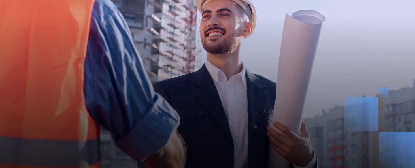 Smiling man in a suit and hard hat holding rolled blueprints shaking hands with a worker in safety vest.