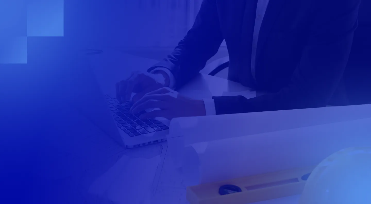 Person in suit typing on a laptop at a desk with architectural plans and a level tool.