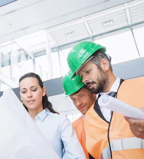 Two men in green hard hats and an orange vest, and a woman, examining architectural plans indoors.