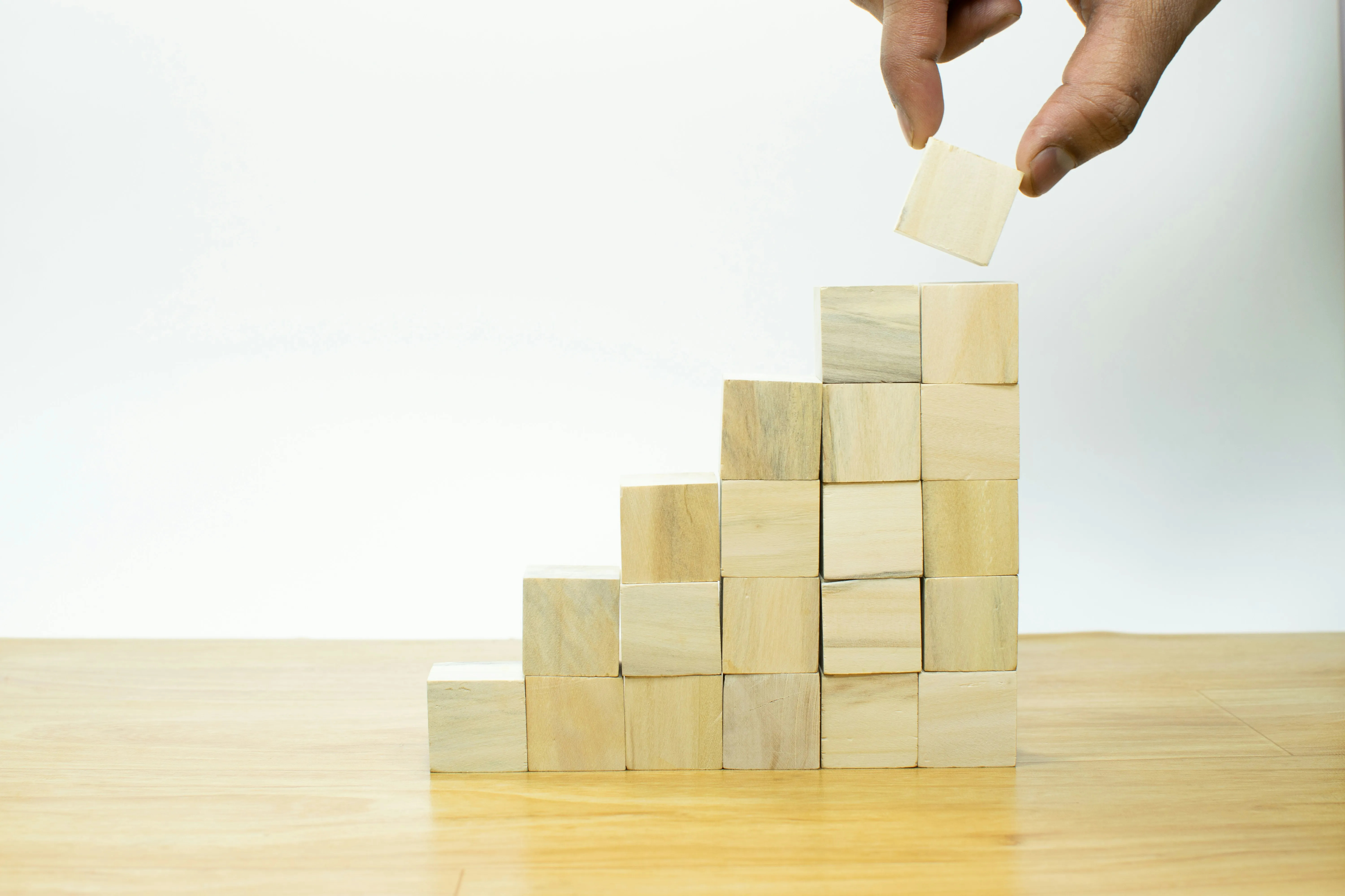 Hand placing the top wooden block on a staircase stack of wooden cubes on a table.