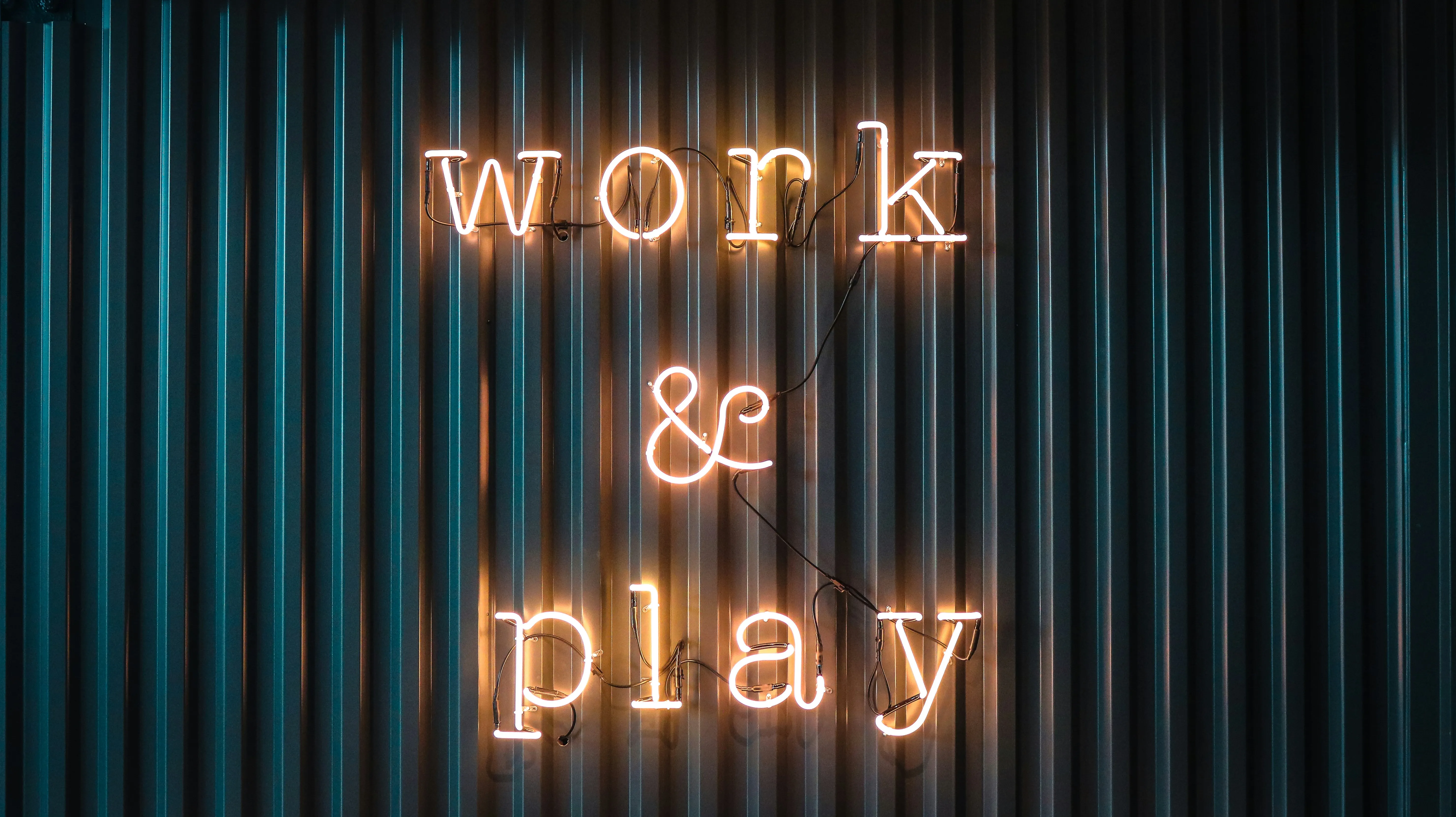 Neon sign on corrugated metal wall displaying the phrase work & play in warm light.