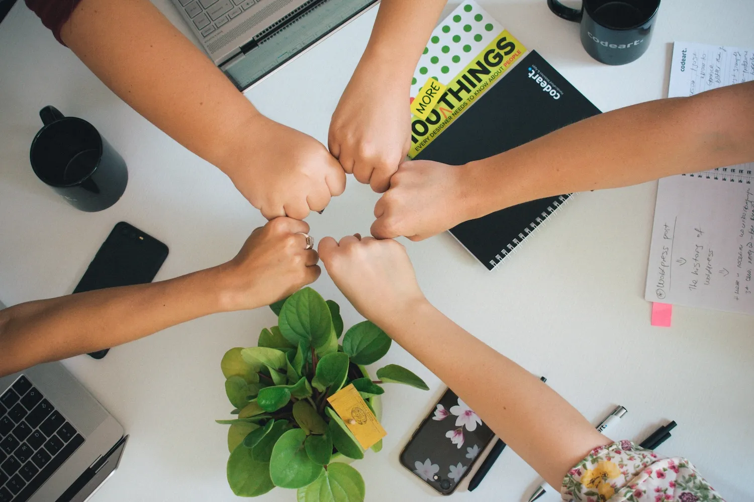 Four fists meeting in the center over a desk with laptops, notebooks, mugs, and a plant.
