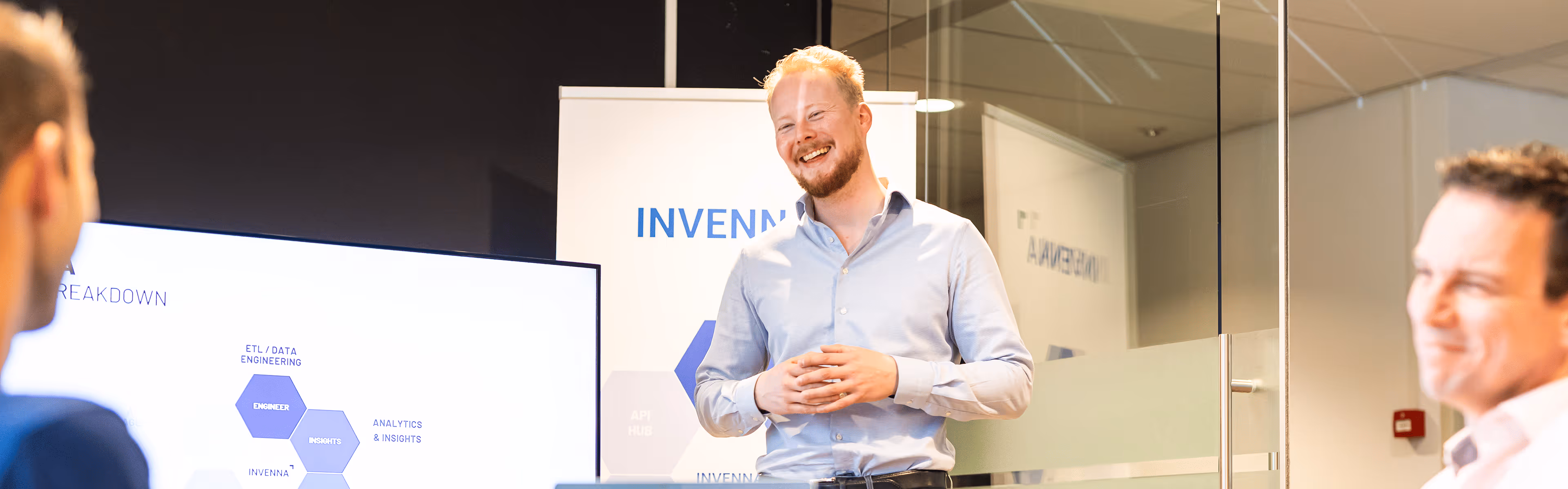 Een lachende man presenteert voor een scherm met een diagram aan een groep collega's in een moderne kantoorruimte.
