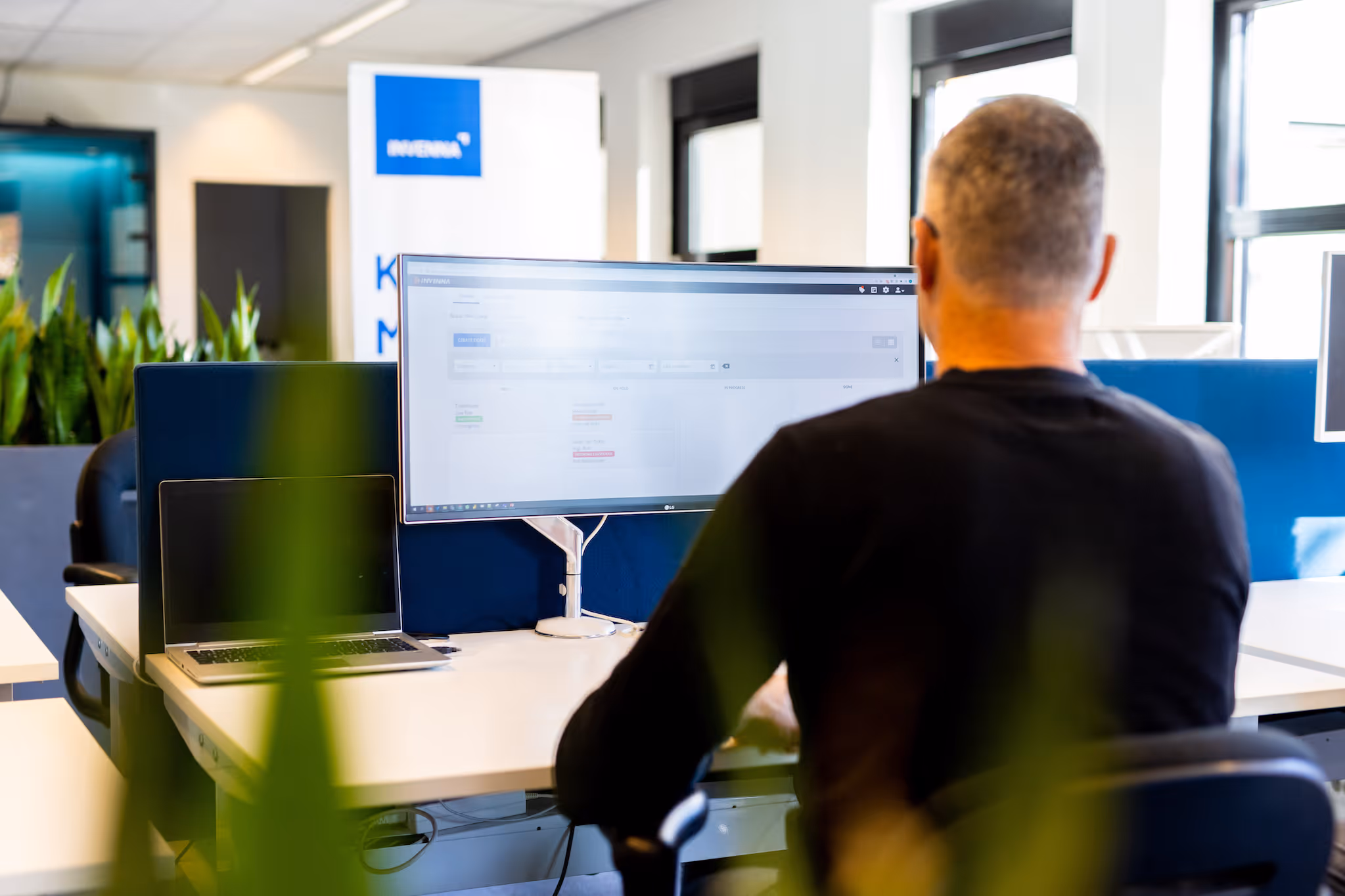 Man werkt op kantoor achter een bureau met laptop en grote monitor in moderne inrichting.
