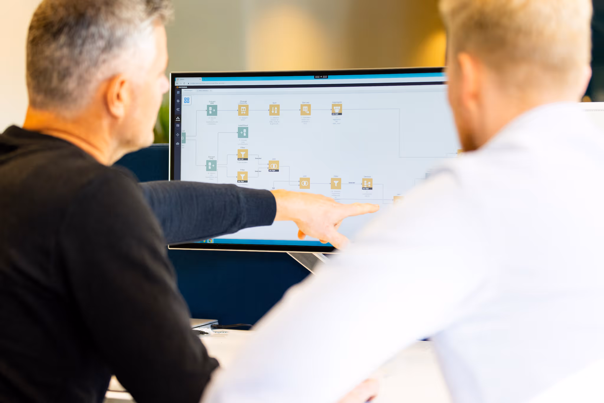 Twee mannen bespreken een stroomdiagram op een computerscherm in een kantooromgeving.