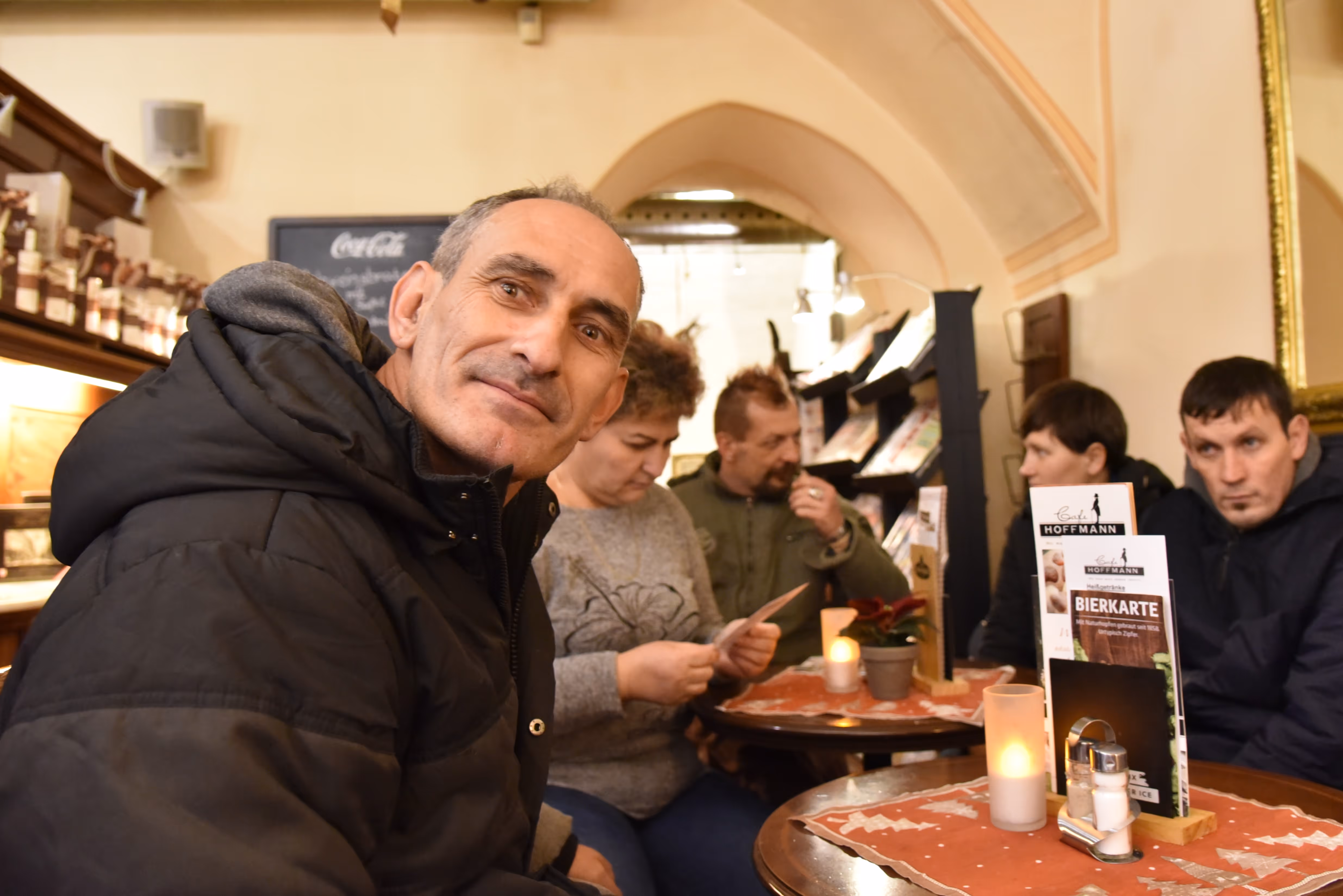 Muškarac u crnoj jakni sjedi za stolom u kafeu s lučnim svodovima i osmjehuje se prema kameri; u pozadini drugi gosti