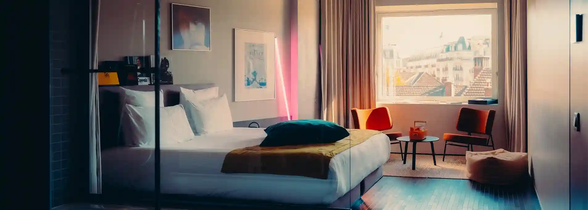 Chambre d'hôtel avec lit double, coussins, chaises orange et grande fenêtre donnant sur des toits urbains.