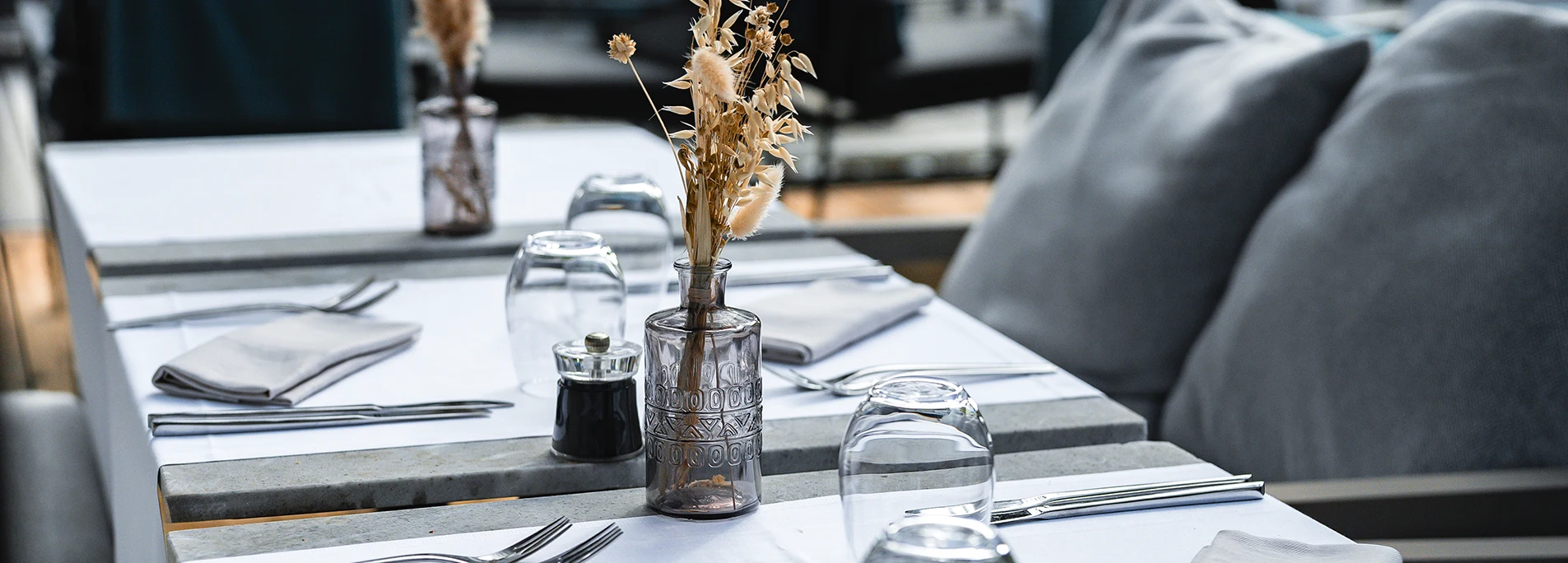 Table de restaurant dressée en blanc avec vase de fleurs séchées, verres à eau et banquette grise en arrière-plan