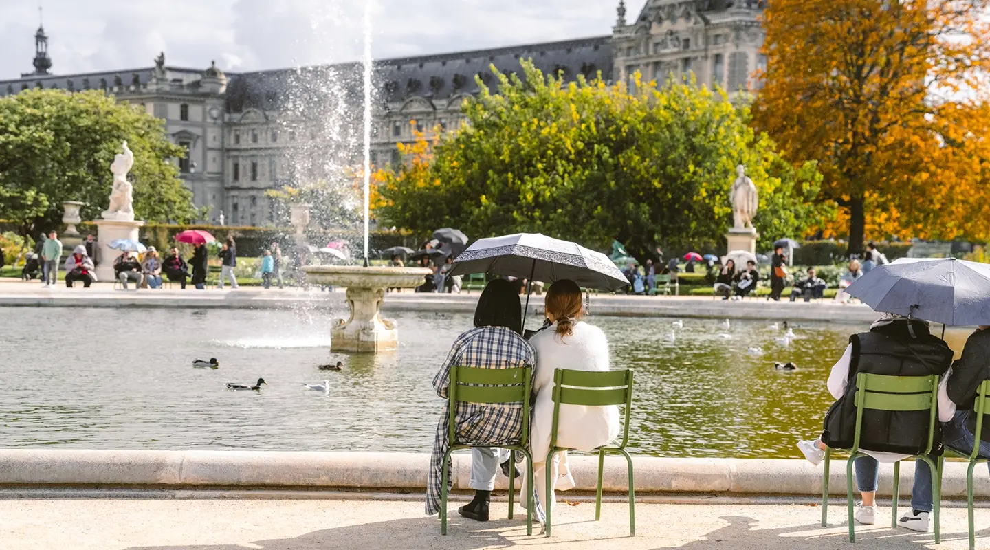 Recadrage vertical resserré sur deux amies partageant un parapluie face au bassin des Tuileries, silhouette du Louvre en fond