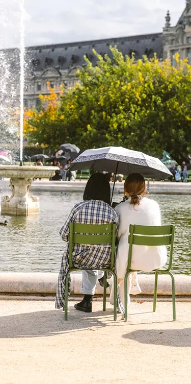 Recadrage vertical resserré sur deux amies partageant un parapluie face au bassin des Tuileries, silhouette du Louvre en fond