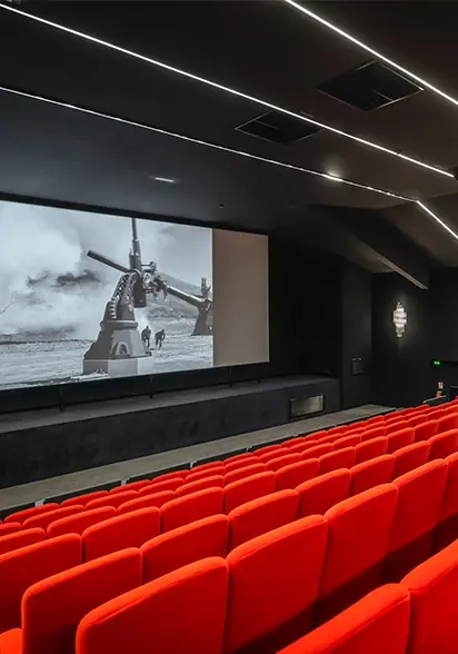 Empty movie theater with rows of red seats, a ceiling with glowing trim, and a screen projecting a black-and-white war film