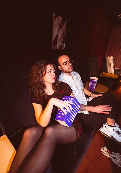 Man and woman sitting in dark cinema seats, holding purple popcorn, absorbed in the movie