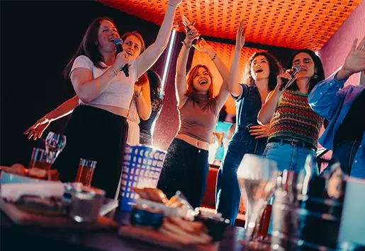A group of young women celebrating and singing karaoke, arms raised around a table filled with drinks and appetizers.