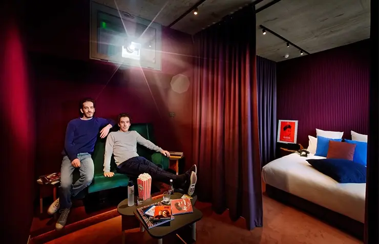 Deux hommes assis sur une banquette verte sous un projecteur de cinéma, chambre attenante aux murs bordeaux cannelés