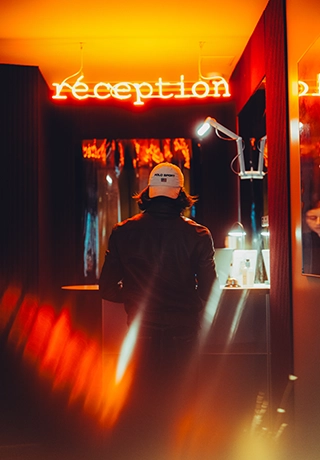 Homme de dos portant une casquette devant la réception d'un hôtel éclairée par un néon rouge.