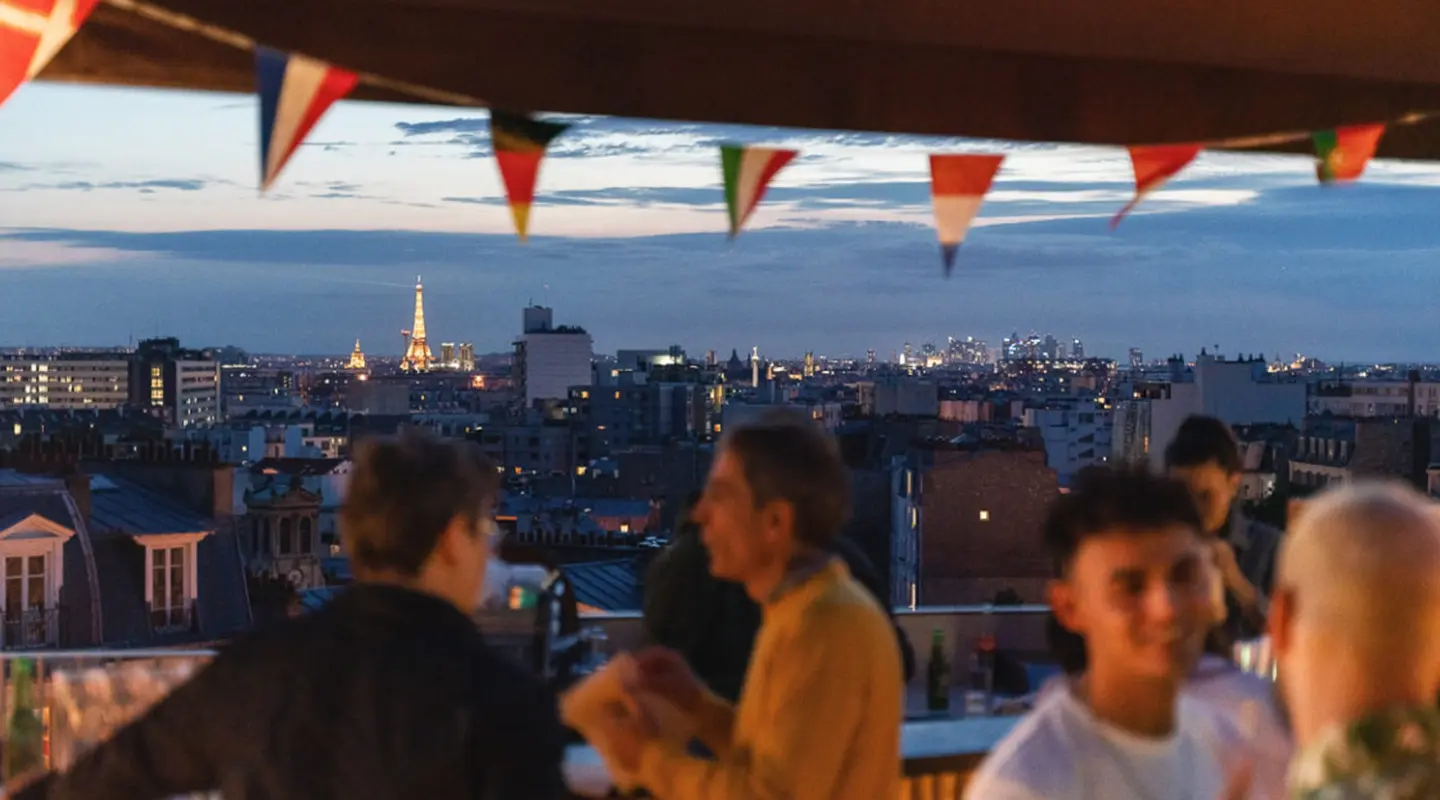 Portrait view of two silhouettes on a rooftop at night, with the sparkling Eiffel Tower on the horizon under colorful bunting