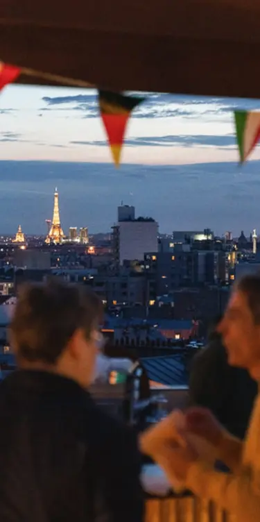 Portrait view of two silhouettes on a rooftop at night, with the sparkling Eiffel Tower on the horizon under colorful bunting