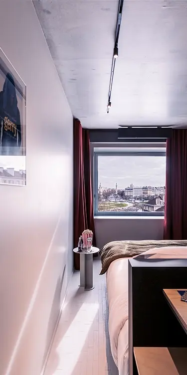 Chambre étroite aux rideaux bordeaux avec fenêtre panoramique sur la ville, veilleuse en forme de pop-corn sur guéridon