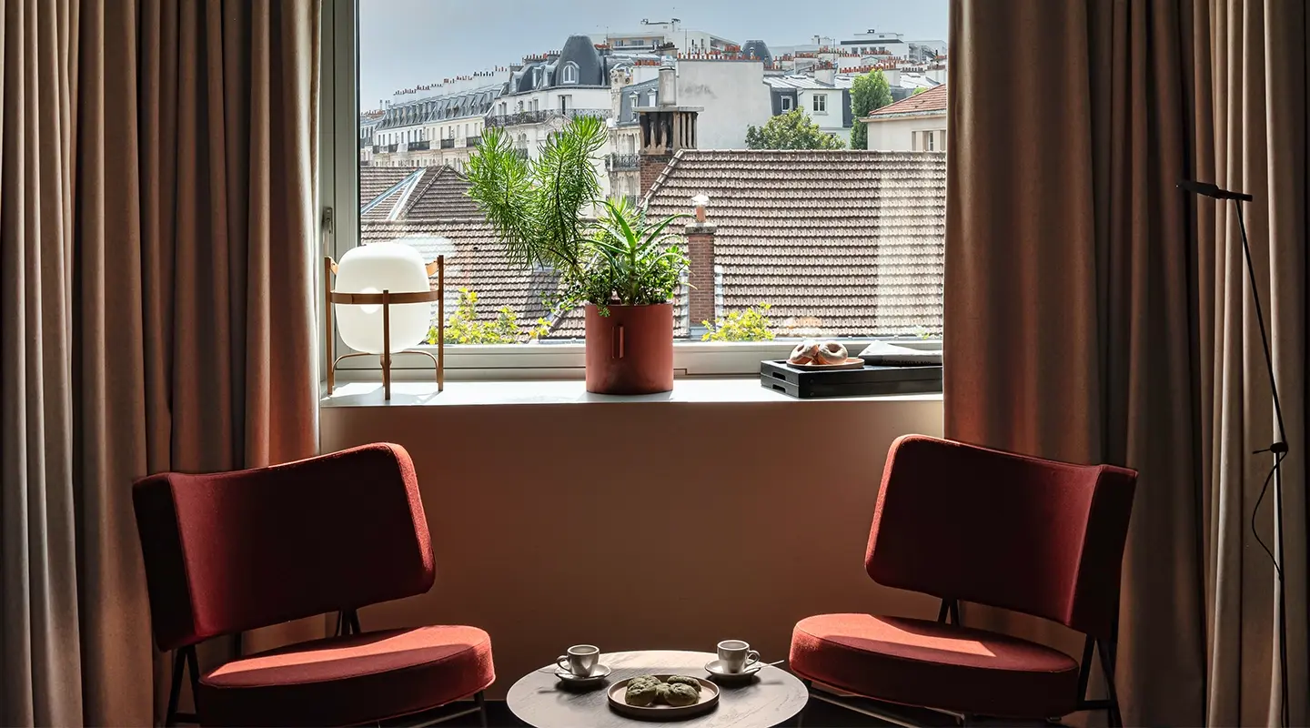 Chambre étroite aux rideaux bordeaux avec fenêtre panoramique sur la ville, veilleuse en forme de pop-corn sur guéridon