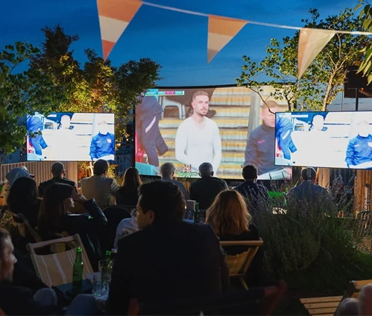Open-air screening on a lush rooftop, with an audience lounging in deckchairs while watching a sporting event broadcast across multiple screens.