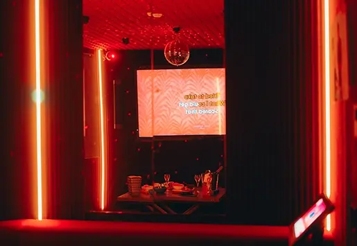 Salle de karaoké privative baignée d'une lumière rouge intense, équipée d'un écran, d'une boule à facettes et d'une table dressée avec des boissons.