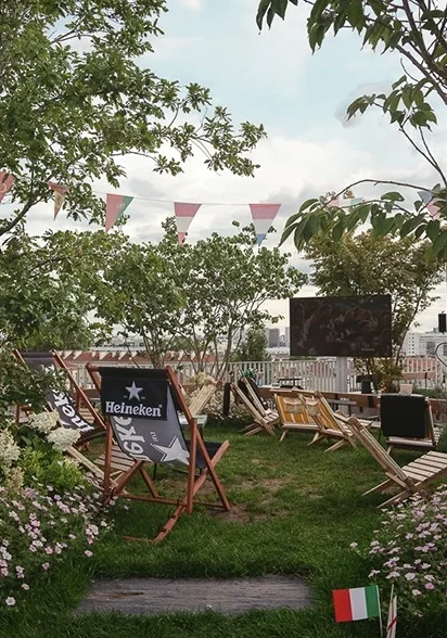 Rooftop garden with Heineken lounge chairs on the grass, a screen showing a football match, and Italian and English flags