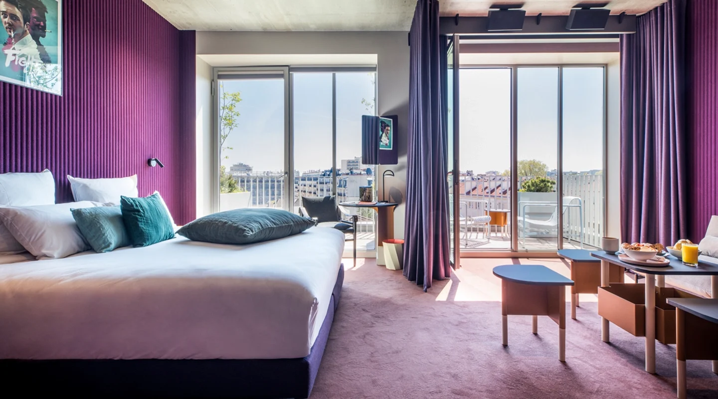 Chambre aux murs cannelés violets, lit à coussins bleu canard et grande baie vitrée ouverte sur un balcon avec vue sur la ville