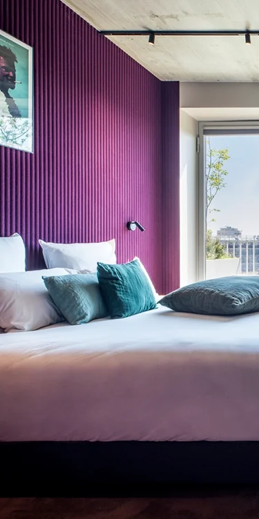 Recadrage vertical resserré sur le lit à coussins bleu canard, mur cannelé violet et porte-fenêtre entrouverte sur un balcon