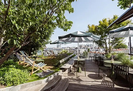 Wooden rooftop terrace with striped lounge chairs, parasols, and green tables, surrounded by lush greenery overlooking the Parisian skyline