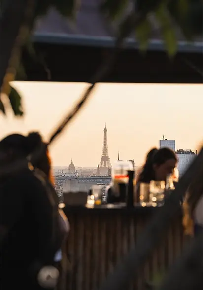 Silhouettes seated at a rooftop bar framing the Eiffel Tower at sunset, with blurred foliage in the foreground