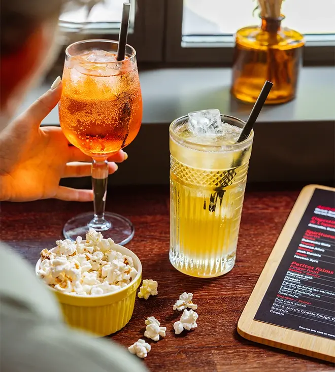 Holding an orange cocktail next to another amber-colored glass, with a bowl of popcorn and a drink menu on a wooden table