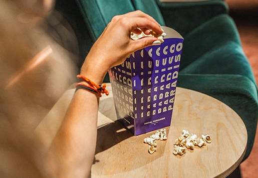 Gros plan sur une main piochant du pop-corn dans une boîte violette « Hôtel Paradiso » posée sur une table en bois.