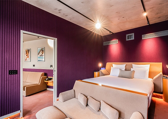 Chambre aux murs cannelés violets et fuchsia, lit à tête de lit orange et vue sur salon attenant aux affiches encadrées