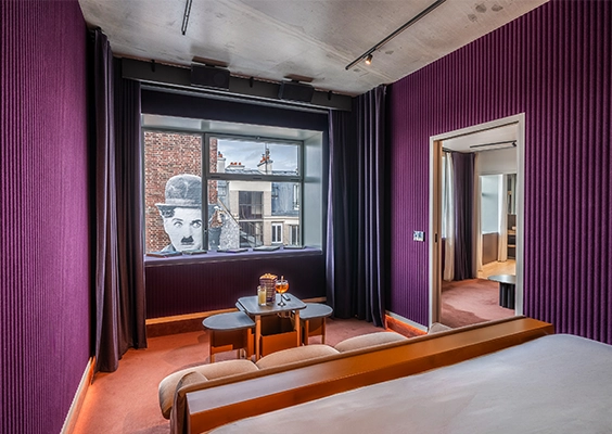 Chambre aux murs cannelés violets, table basse ronde et guéridons devant une fenêtre encadrée de rideaux gris, fresque murale visible en arrière-plan