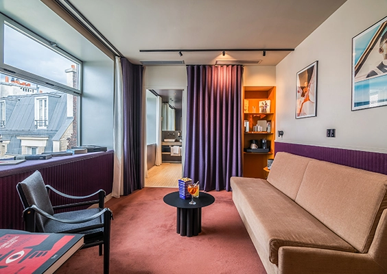 Salon avec canapé camel et fauteuil noir, table basse ronde et rideau violet ouvrant sur une salle de bain, vue sur les toits parisiens