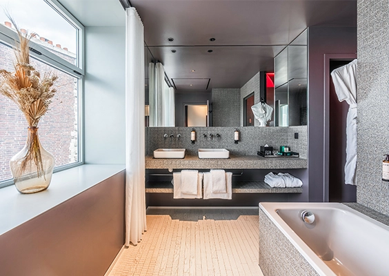 Salle de bain carrelée en mosaïque grise avec double vasque, grand miroir et baignoire encastrée près d'une fenêtre à bouquet de fleurs séchées