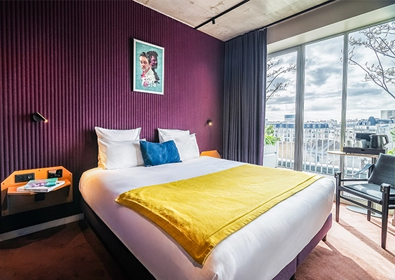 Chambre d'hôtel lumineuse avec mur violet, lit double et baies vitrées sur la ville.