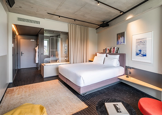 Chambre d'hôtel contemporaine avec lit double, tapis beige, sol en carrelage sombre et salle de bains semi-vitrée.