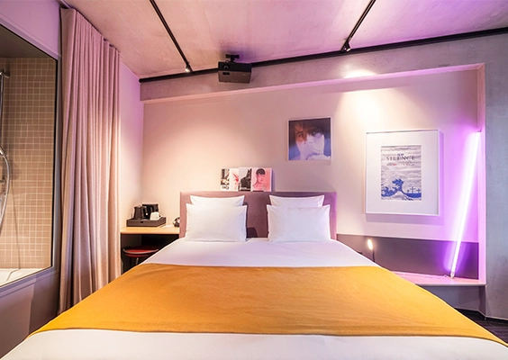 Chambre d'hôtel moderne avec lit double au couvre-lit jaune, éclairage néon violet et rétroprojecteur au plafond.