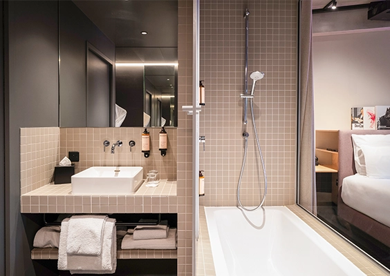 Vue sur une salle d'eau design carrelée de beige clair, attenante à une chambre, comprenant une vasque carrée sur plan et une baignoire.