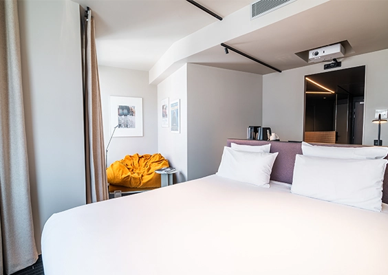Chambre d'hôtel moderne et lumineuse dotée d'un grand lit double aux draps blancs, d'un fauteuil pouf jaune moutarde au fond et d'un rétroprojecteur fixé au plafond.