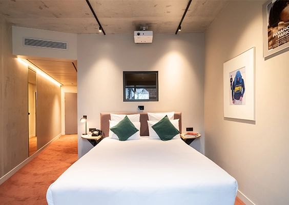 Chambre d'hôtel moderne avec lit double, coussins verts et vidéo-projecteur au plafond.
