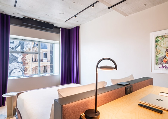 Chambre d'hôtel moderne avec lit double, coin bureau intégré en tête de lit et rideaux violets près d'une fenêtre donnant sur la ville.