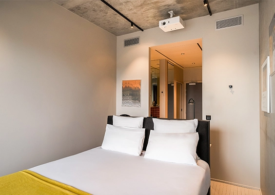 Chambre d'hôtel épurée avec lit double, draps blancs, plaid jaune et projecteur fixé au plafond.