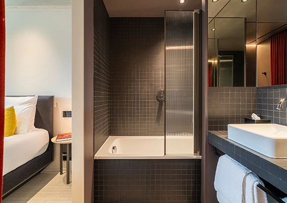 Salle de bains moderne en carrelage sombre avec baignoire, pare-douche et lavabo, communiquant avec la chambre.