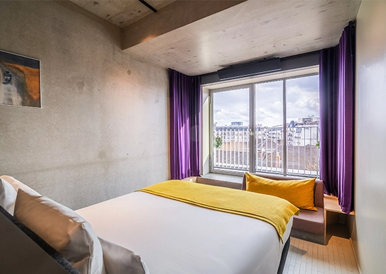 Chambre d'hôtel moderne avec lit double au plaid jaune, mur en béton brut et grande fenêtre encadrée de rideaux violets.