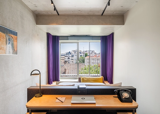 Vue axiale sur un espace bureau en bois avec ordinateur et téléphone vintage, ouvrant sur la chambre et une grande fenêtre encadrée de rideaux violets.